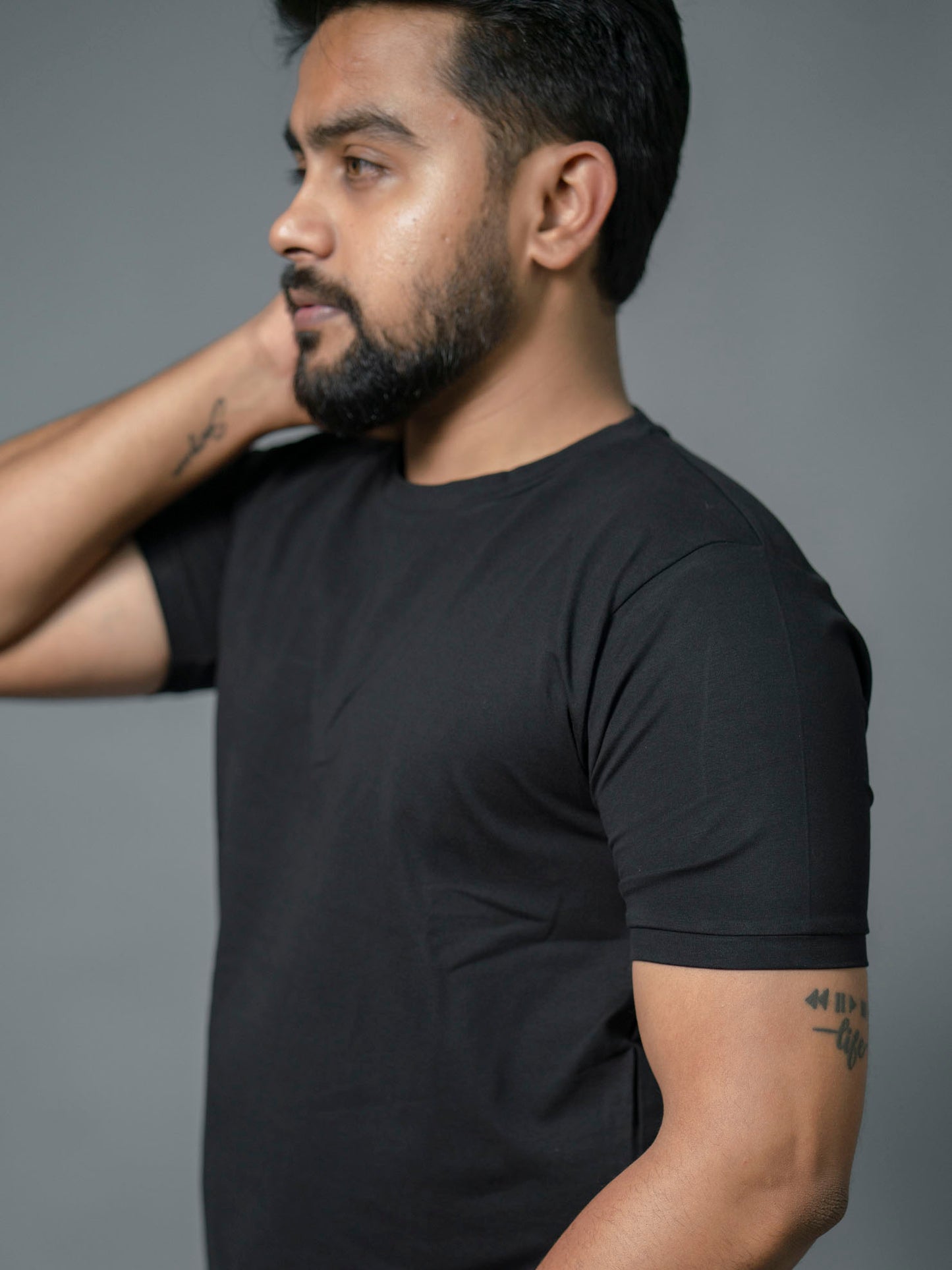 Man in a side pose wearing a black "The No Name Club" brand crew/round neck t-shirt against a gray background.