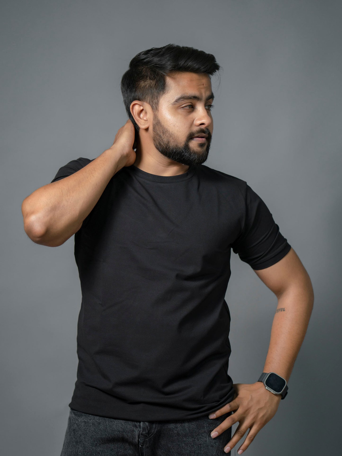 Man wearing a black "The No Name Club" brand crew/round  neck t-shirt against a gray background. One Hand on Neck, the other on the waist.